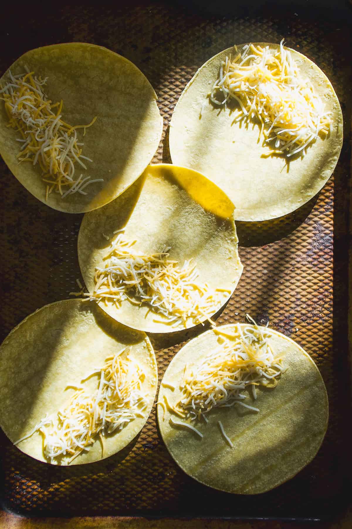 Tortillas spread out on a baking sheet with cheese on top about to be baked.