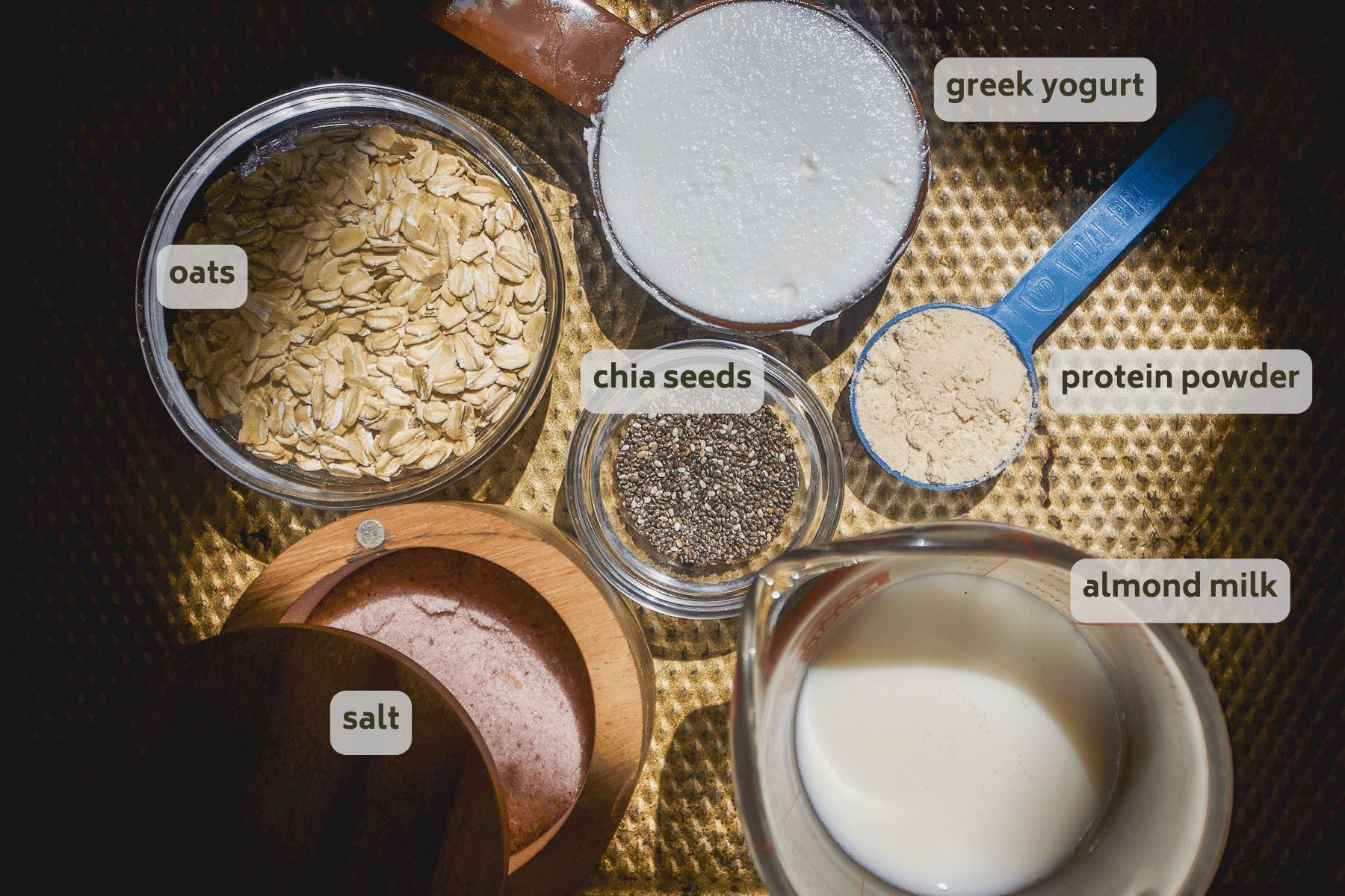 Top-down view of ingredients for high protein overnight oats: oats, chia seeds, salt, Greek yogurt, protein powder, and almond milk. Each is in a separate, labeled container and arranged on a textured surface.