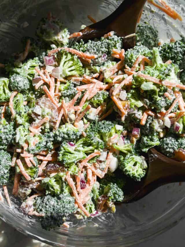 A close-up of broccoli salad in a glass bowl, mixed with shredded carrots, chopped red onion, and a creamy dressing, with wooden salad tongs partially visible.
