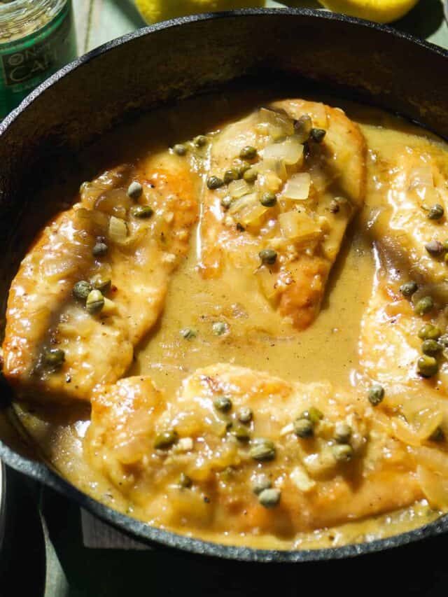 Chicken piccata in a cast iron skillet with capers on top in a lemon sauce.