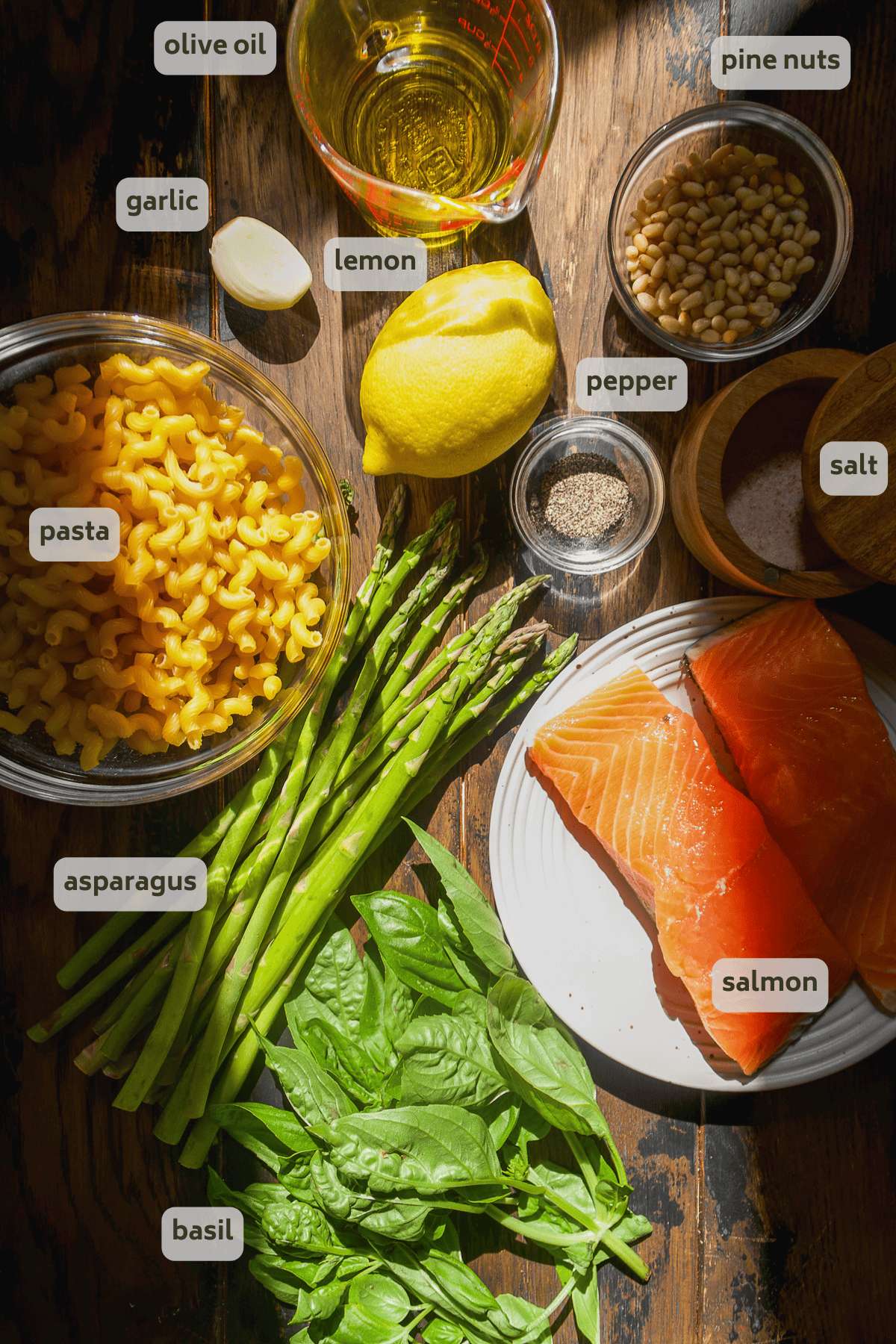 Pesto salmon pasta ingredients on a wooden board with labels.