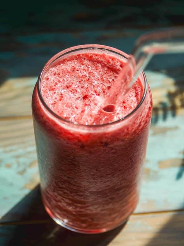 A glass of red blended fruit smoothie with a clear straw, photographed from above, sits on a sunlit wooden surface.