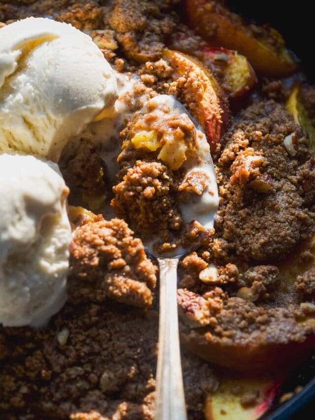 A close-up of a baked peach crisp topped with two scoops of vanilla ice cream, with a spoon scooping out a portion, showing the crumbly topping over sliced peaches.