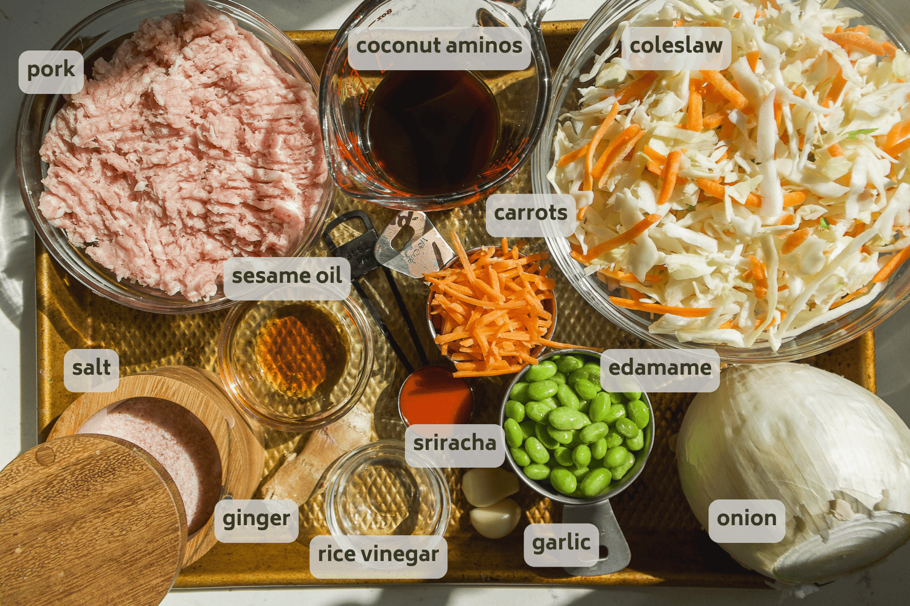 A tray holds ingredients for a healthy egg roll in a bowl with coleslaw mix: ground pork, coconut aminos, carrots, edamame, onion, garlic, ginger, rice vinegar, sriracha, sesame oil, and salt—all clearly labeled.