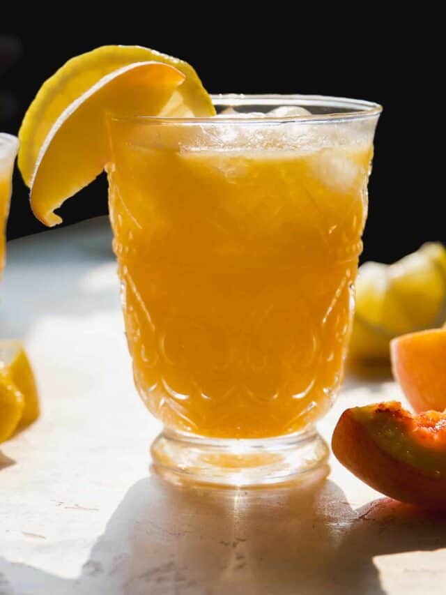 A glass of iced orange-colored drink garnished with a lemon wedge sits on a light surface with lemon and peach slices in the background, illuminated by natural sunlight.