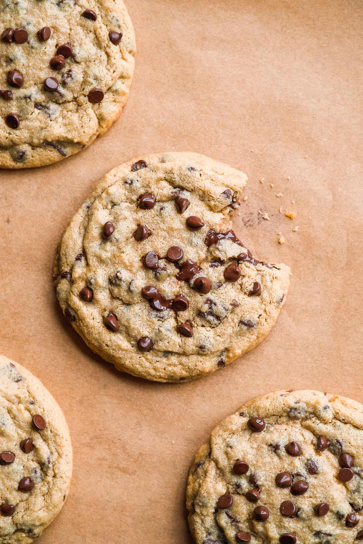 Four homemade chocolate chip cookies on a piece of brown parchment paper. One cookie has a small bite taken out of it.