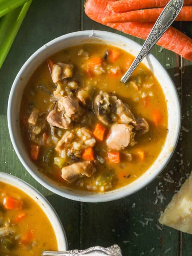 A bowl of hearty chicken and rice soup with diced carrots and celery, placed on a green surface next to fresh carrots and a piece of Parmesan cheese. A spoon rests inside the bowl.
