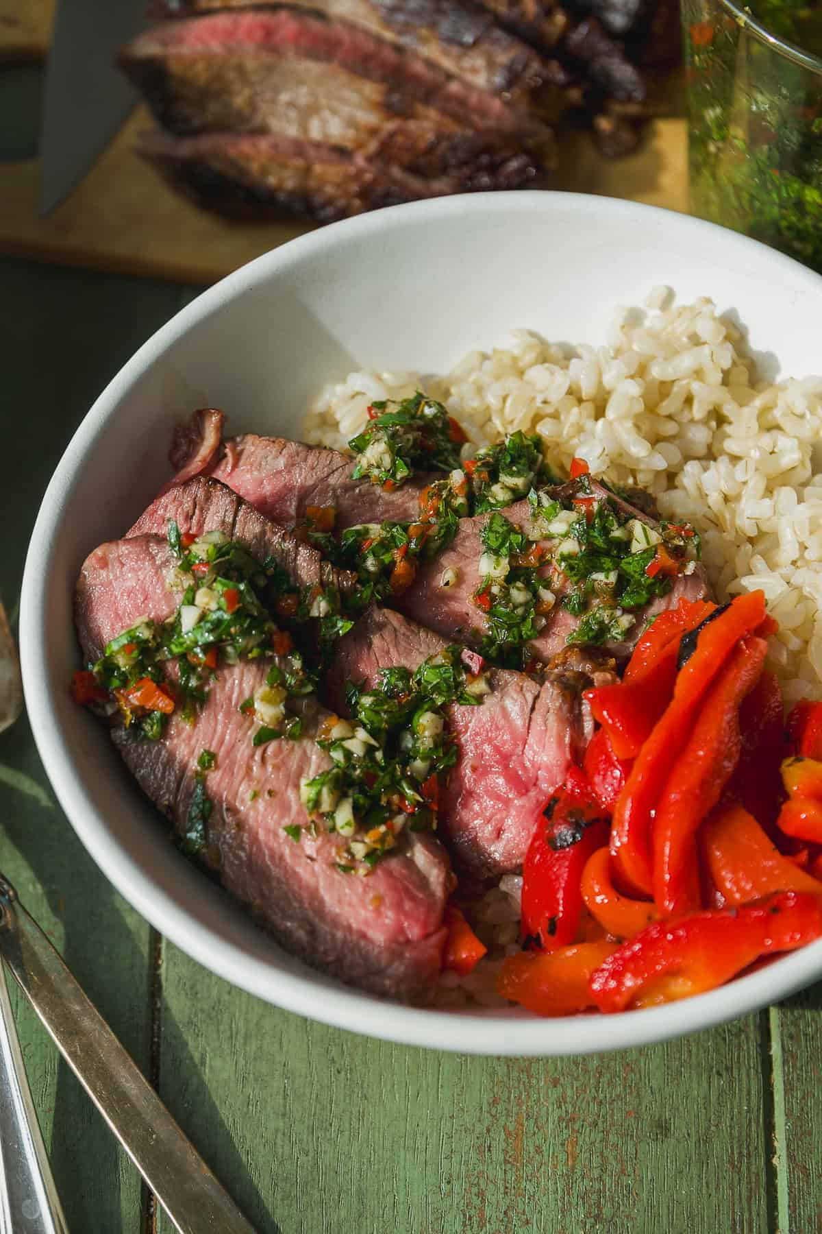 Steak with chimichurri sauce on top in a bowl with rice and roasted red peppers.