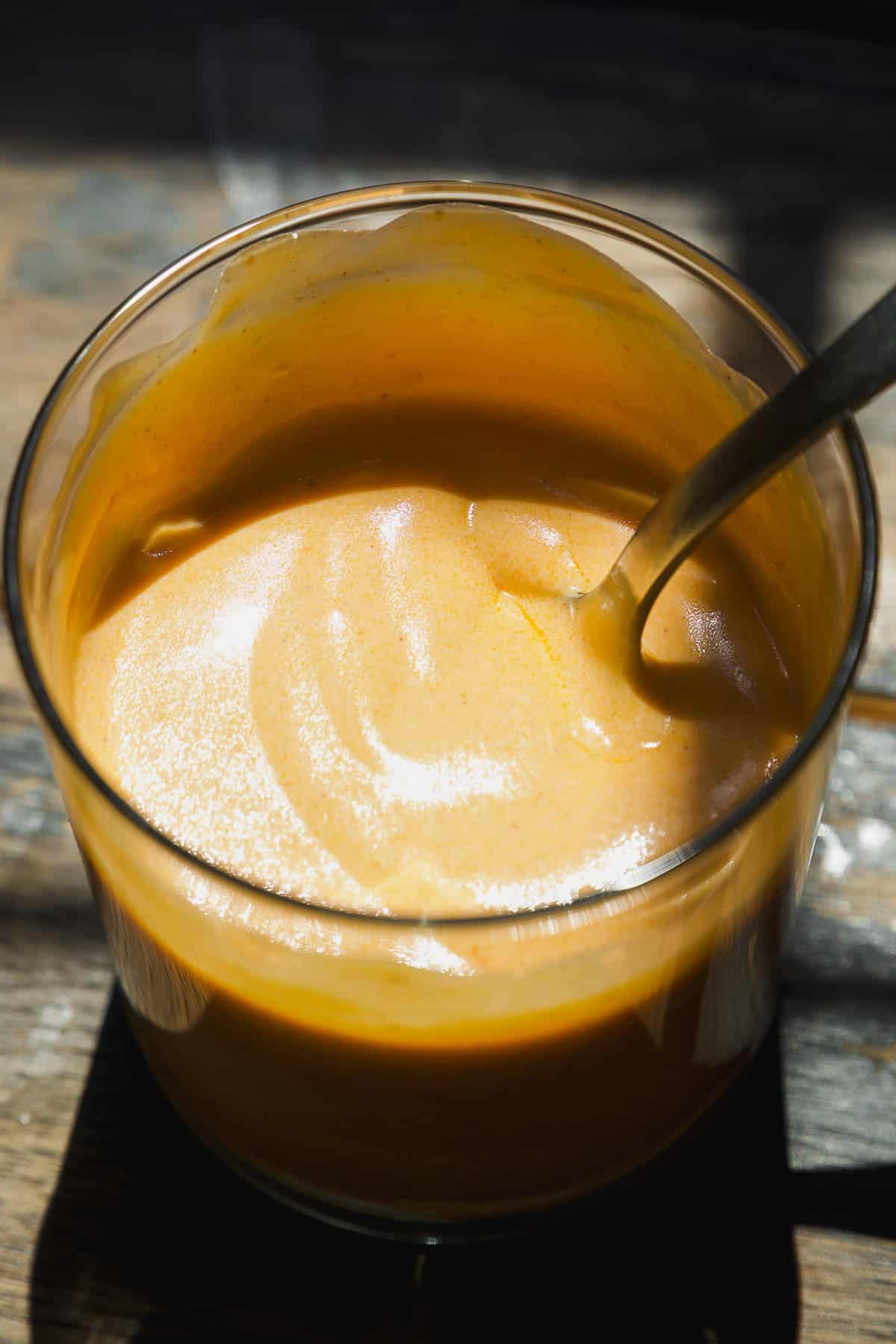 A close-up of a glass filled with creamy orange sauce or dip, with a metal spoon partially submerged in it. Sunlight casts shadows and highlights on the surface of the sauce.