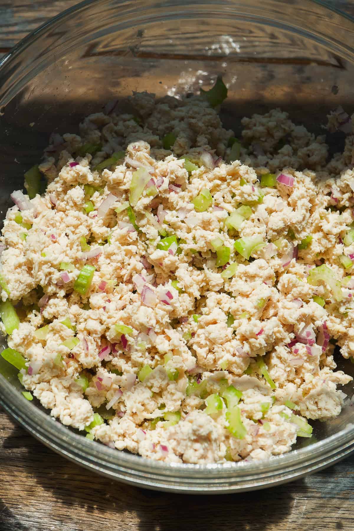 A glass bowl filled with a mixture of chopped chicken, diced celery, and red onion, sitting on a wooden surface in bright natural light.