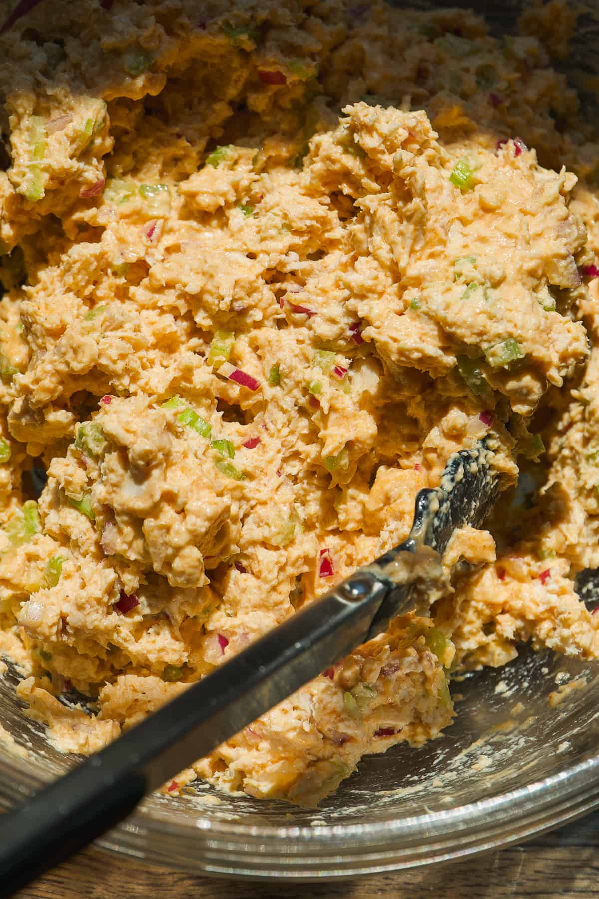 A close-up of a glass bowl filled with a chunky, orange-tinted mixture containing visible pieces of red onion and green vegetables. A black spoon rests inside the bowl.