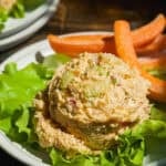 A scoop of chicken salad with diced celery and onions is served on a bed of lettuce, accompanied by carrot sticks on a white plate. The dish is set on a wooden table in natural light.