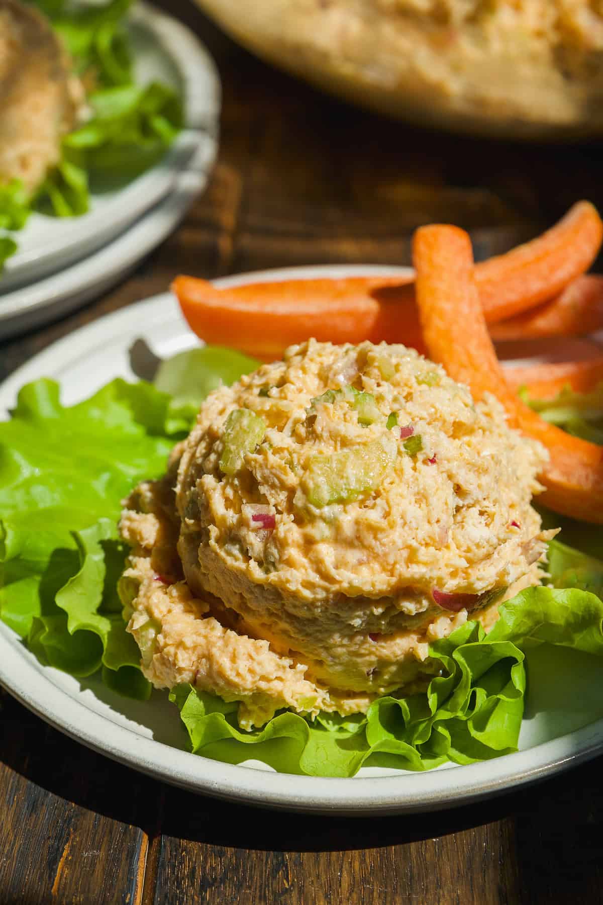A scoop of chicken salad with diced celery and onions is served on a bed of lettuce, accompanied by carrot sticks on a white plate. The dish is set on a wooden table in natural light.