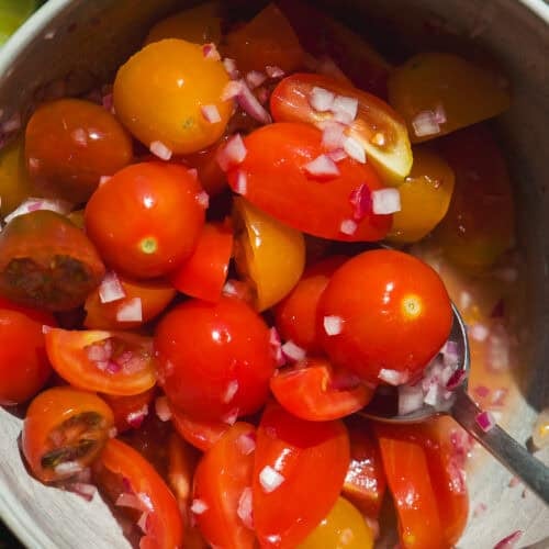 A bowl of cherry tomatoes, both red and yellow, mixed with finely chopped red onions—perfect as a healthy burger bowl topping. A metal spoon rests in the bowl, with sliced lime visible at the edge of the photo.