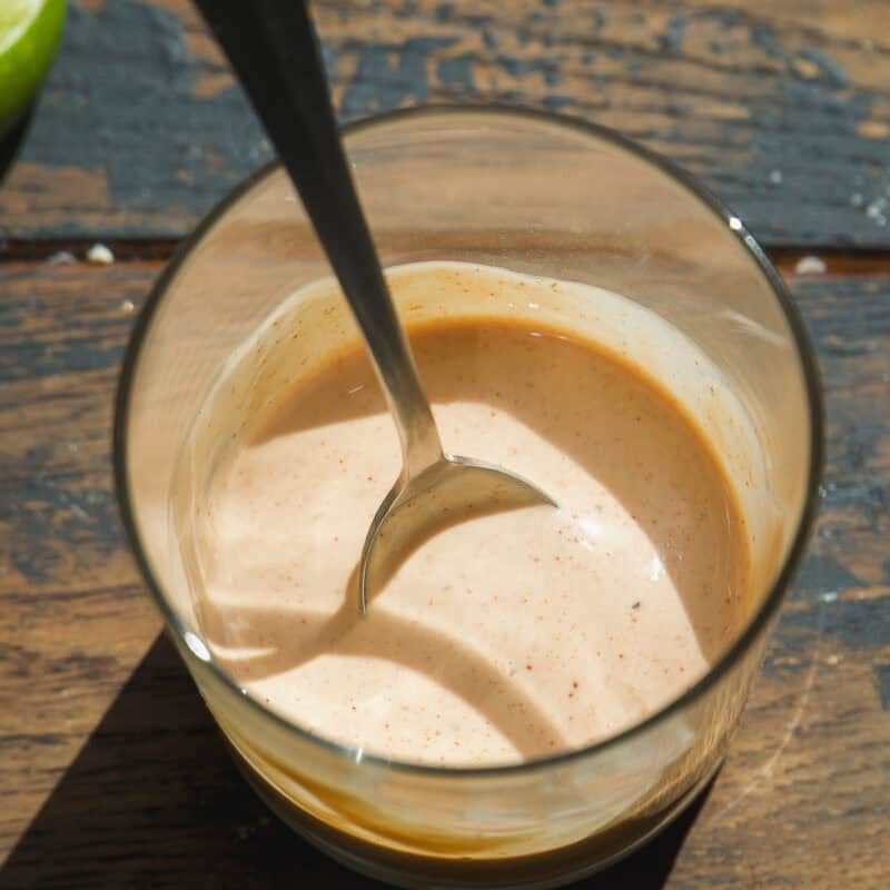 A glass filled with a creamy, light orange sauce, perfect for drizzling over a healthy burger bowl, sits on a wooden surface. A metal spoon rests inside, and a sliced lime is partially visible in the upper left corner.