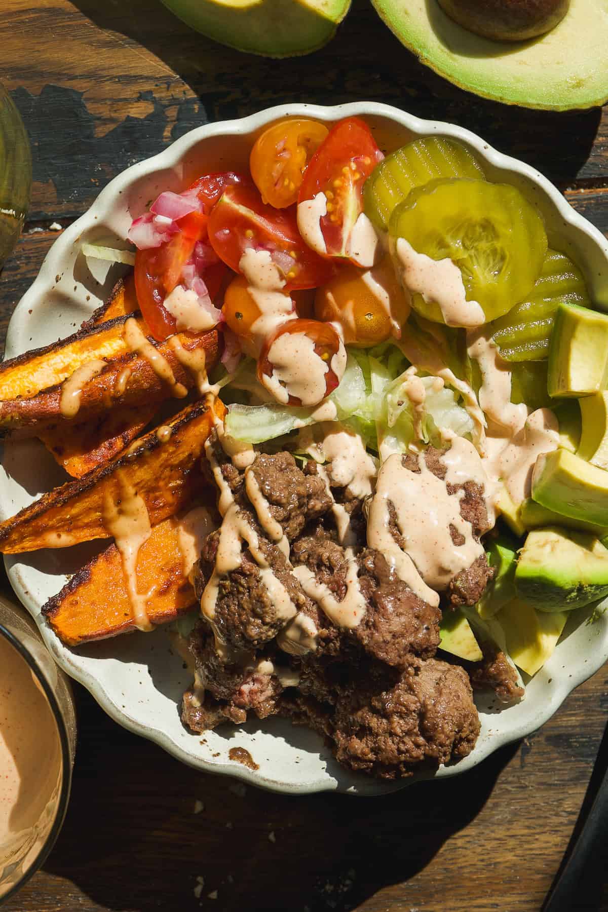 A healthy burger bowl filled with ground beef, sliced avocado, sweet potato wedges, lettuce, tomato, and pickles, all drizzled with creamy sauce and artfully arranged on a wooden table.
