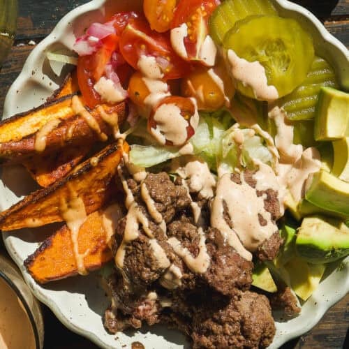 A healthy burger bowl filled with ground beef, sliced sweet potatoes, avocado chunks, pickle slices, lettuce, cherry tomatoes, and a drizzle of creamy sauce sits on a wooden table with halved avocados nearby.