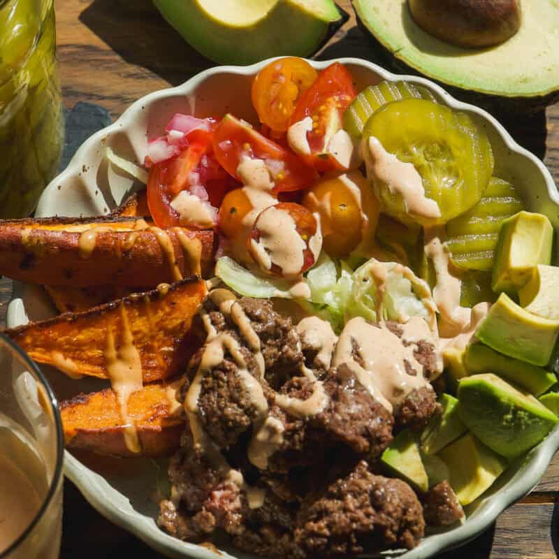 A healthy burger bowl featuring ground beef, sweet potato wedges, sliced avocado, lettuce, tomato, pickles, and creamy dressing. Sliced avocado, limes, and a glass of dressing sit on the wooden table nearby.