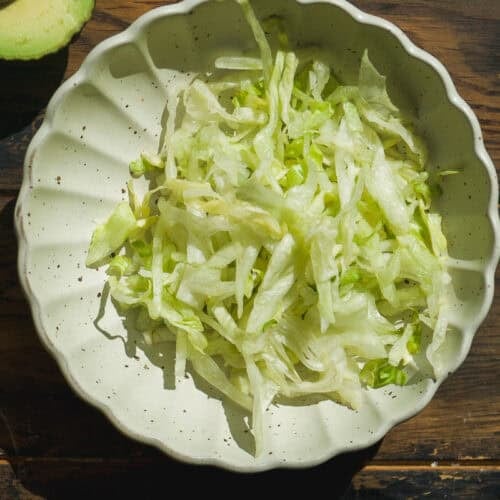 A scalloped ceramic bowl filled with shredded iceberg lettuce sits on a wooden table—perfect as a base for your healthy burger bowl. Part of an avocado and a lime are visible in the corner of the image.
