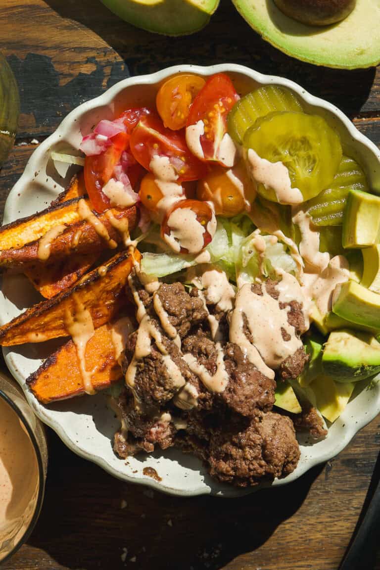 A healthy burger bowl filled with ground beef, sliced sweet potatoes, avocado chunks, pickle slices, lettuce, cherry tomatoes, and a drizzle of creamy sauce sits on a wooden table with halved avocados nearby.