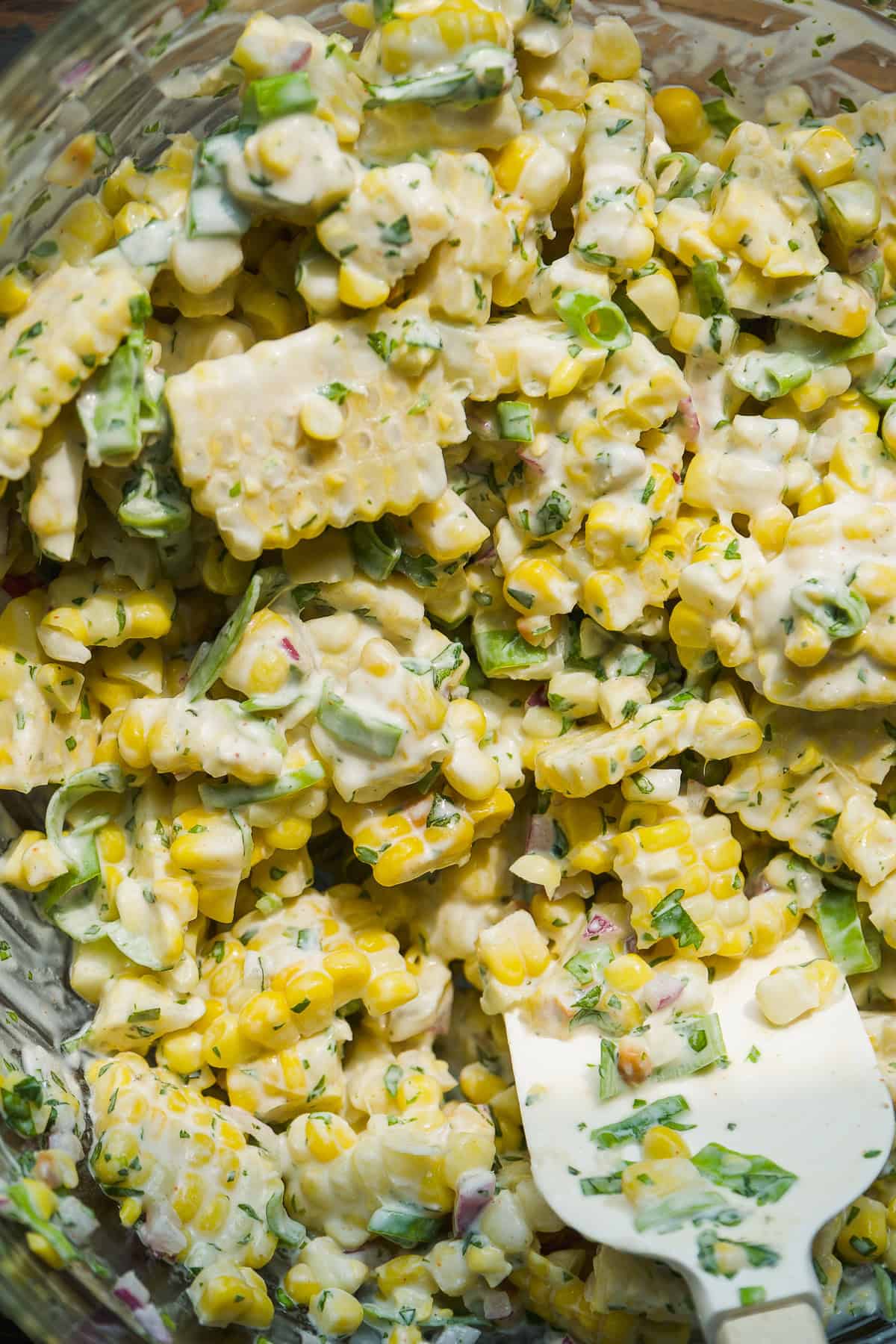 Close-up of a creamy corn salad with chopped green onions, herbs, and diced red onions, mixed together with a white spatula in a glass bowl.