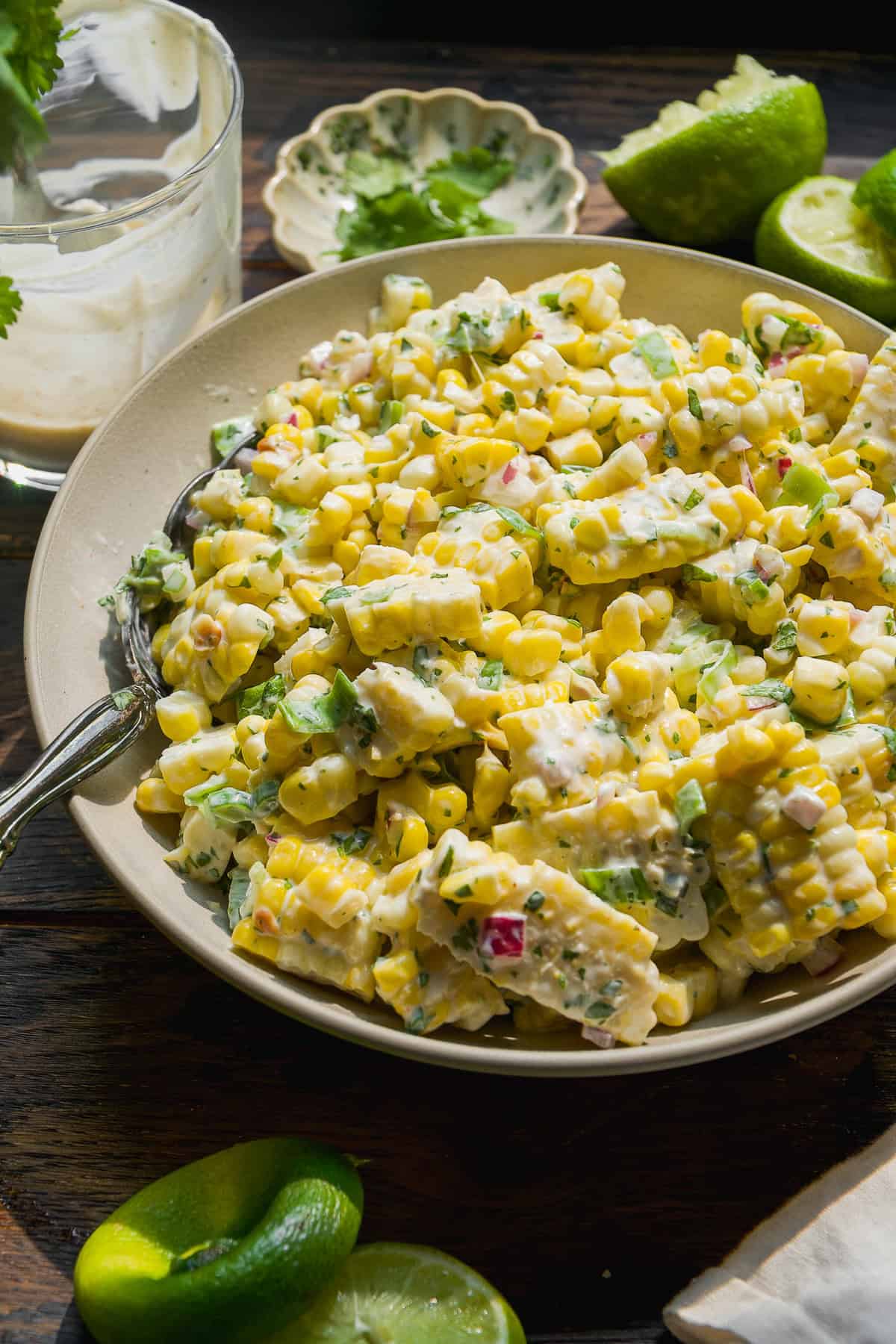 A bowl of creamy corn salad with chopped vegetables, herbs, and a light dressing, served with a spoon. Lime wedges and a glass with leftover dressing are nearby on a rustic wooden table.