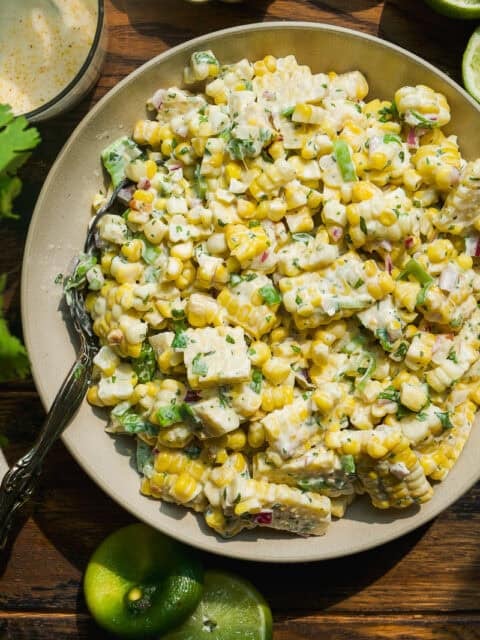 A bowl of creamy corn salad with diced avocado, chopped herbs, and lime wedges on the side, served on a wooden table with a fork in the bowl—an easy corn salad recipe perfect for any meal.