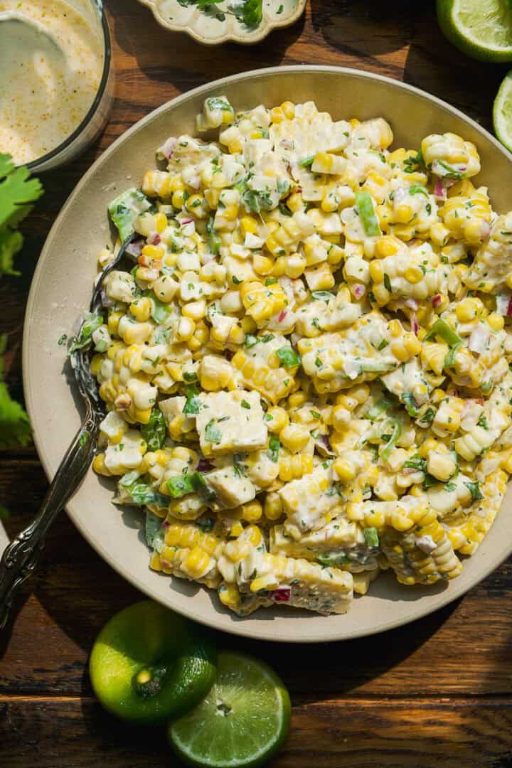 A bowl of creamy corn salad with diced avocado, chopped herbs, and lime wedges on the side, served on a wooden table with a fork in the bowl—an easy corn salad recipe perfect for any meal.