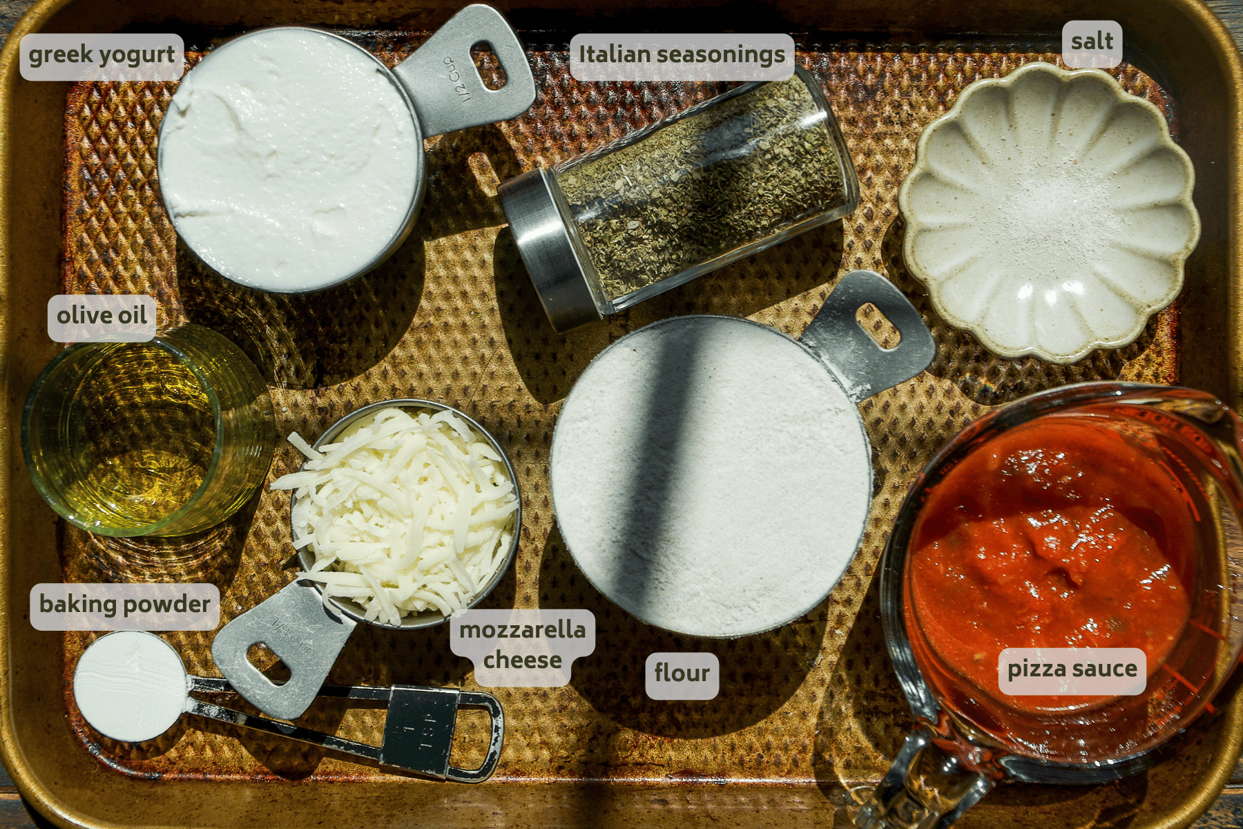 A baking tray displays measured ingredients for greek yogurt pizza dough: Greek yogurt, olive oil, baking powder, mozzarella cheese, Italian seasonings, flour, salt, and a jug of pizza sauce—each labeled for this delicious greek yogurt pizza dough recipe.