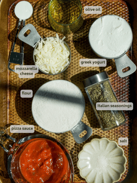A baking tray holds labeled ingredients in bowls and measuring cups: baking powder, olive oil, shredded mozzarella cheese, Greek yogurt, flour, Italian seasonings, pizza sauce, and salt.