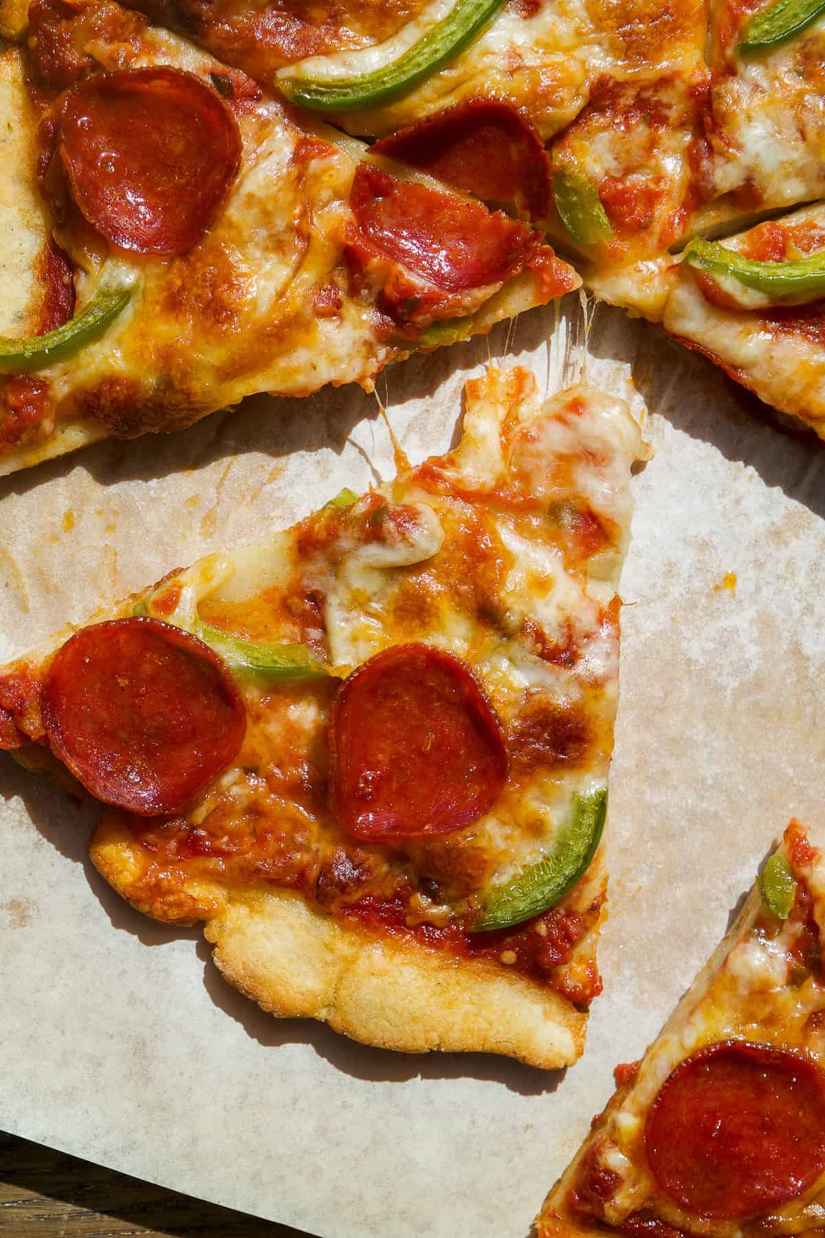 A slice of pizza with pepperoni, green bell pepper, and melted cheese sits on parchment paper, with other pizza slices partially visible around it. The crust is golden brown and the cheese is slightly browned.