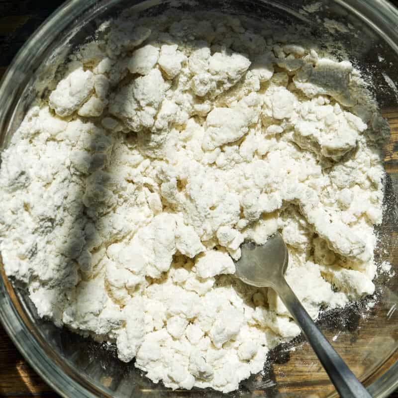 A glass bowl containing clumpy flour mixture with a metal fork inside, set on a wooden surface with sunlight casting a shadow across part of the bowl.