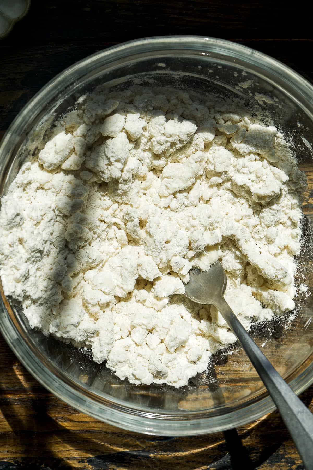 A glass bowl containing clumpy flour mixture with a metal fork inside, set on a wooden surface with sunlight casting a shadow across part of the bowl.