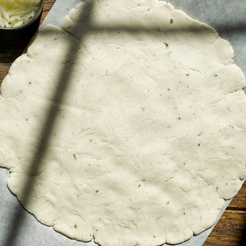 A round, uncooked pizza dough is rolled out on parchment paper on a wooden surface, with shadows crossing over it. A small bowl of shredded cheese sits in the upper left corner.