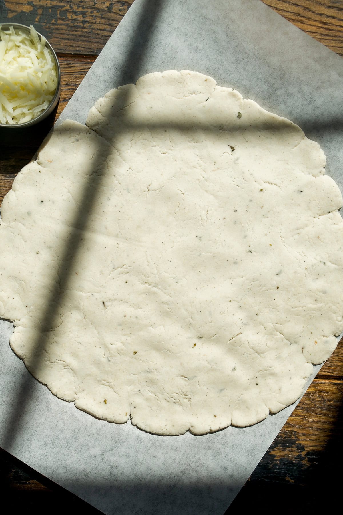 A round, uncooked pizza dough is rolled out on parchment paper on a wooden surface, with shadows crossing over it. A small bowl of shredded cheese sits in the upper left corner.