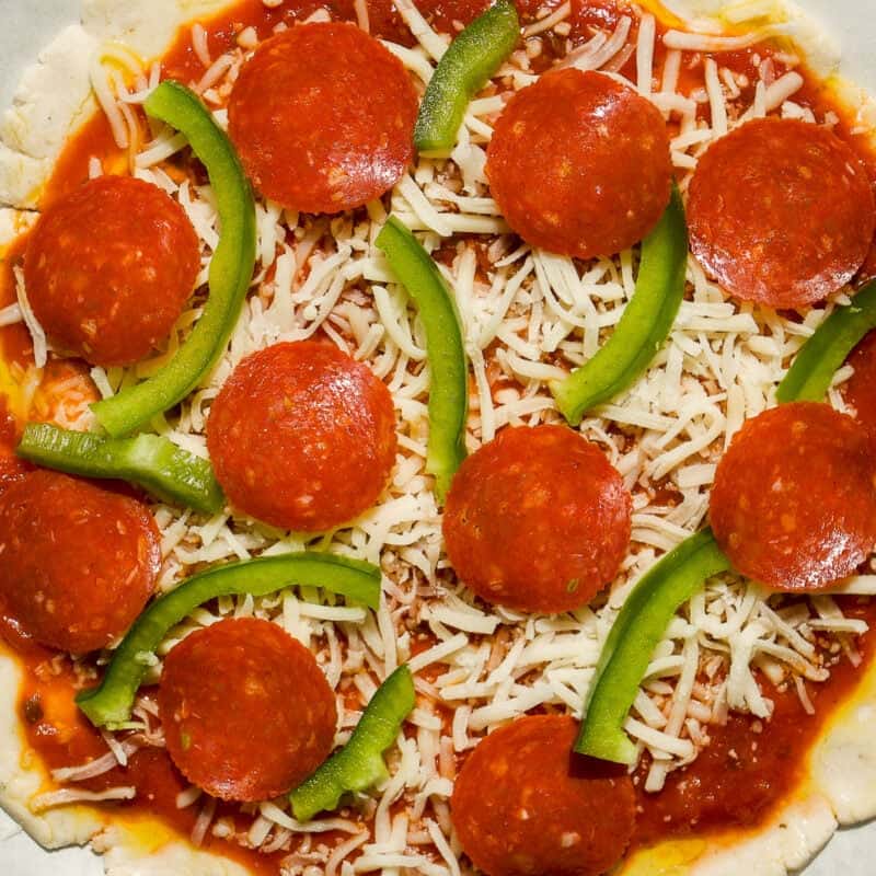 Uncooked pizza with tomato sauce, shredded mozzarella cheese, pepperoni slices, and green bell pepper strips on a round crust, placed on parchment paper.