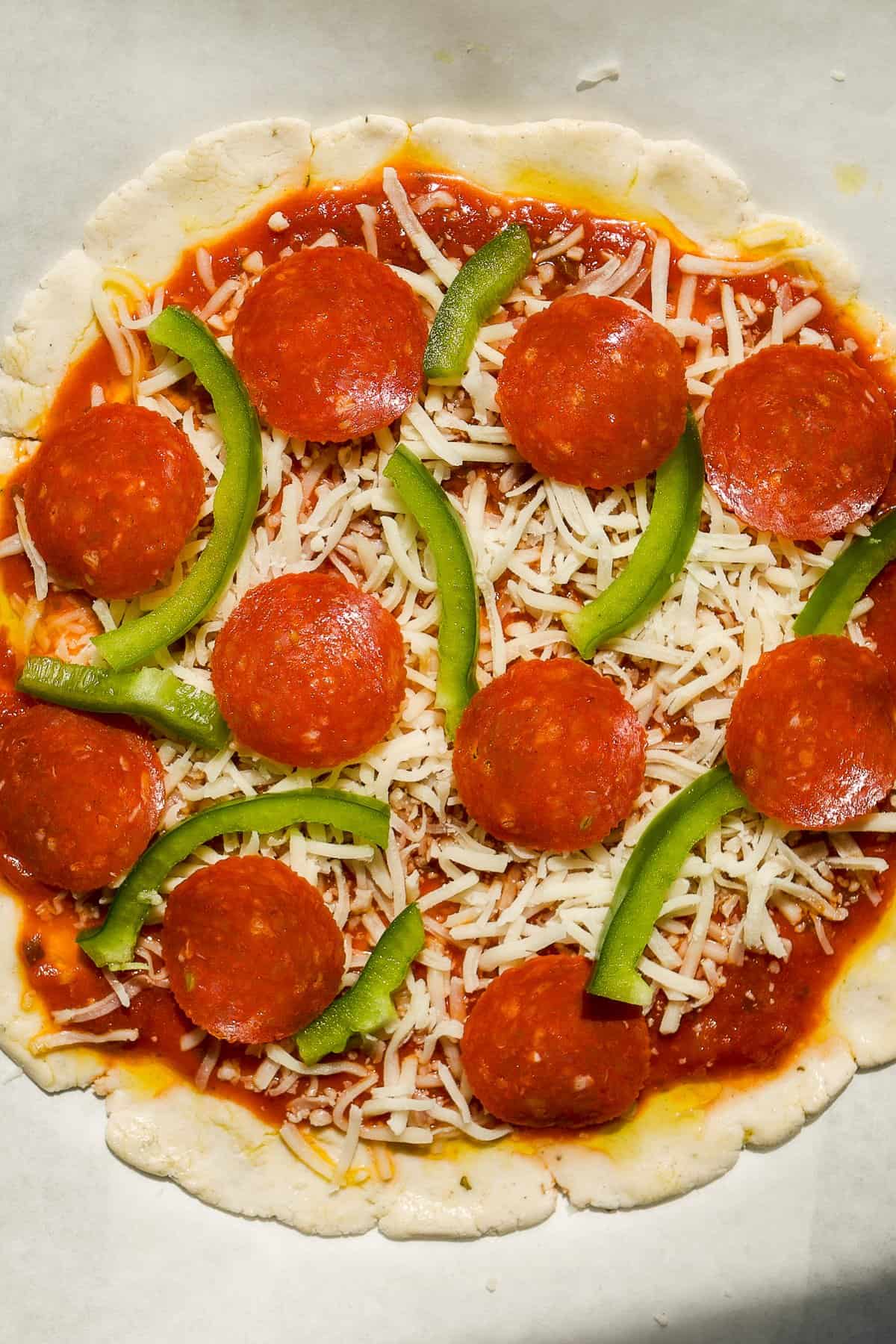 Uncooked pizza with tomato sauce, shredded mozzarella cheese, pepperoni slices, and green bell pepper strips on a round crust, placed on parchment paper.
