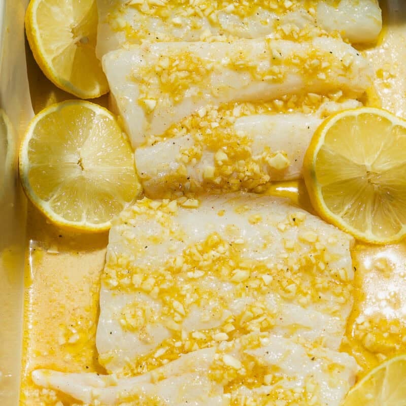 Raw fish fillets marinated in a yellow garlic and lemon sauce, topped with minced garlic, and surrounded by halved lemons in a rectangular dish.