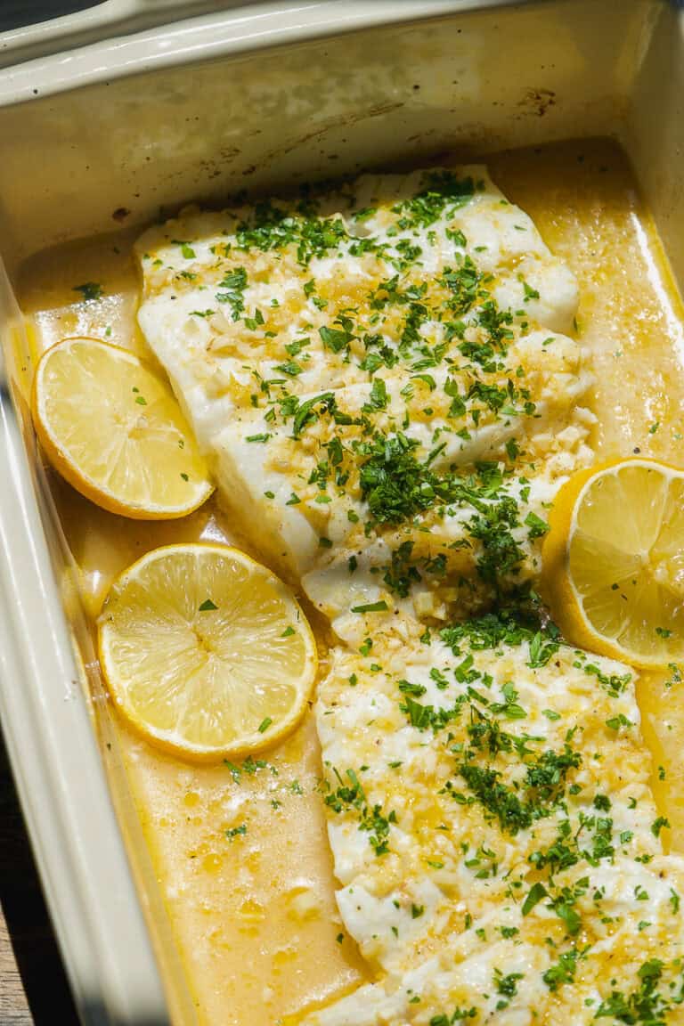 Two baked cod fillets in a baking dish, topped with chopped herbs and garlic, surrounded by lemon slices and a light buttery sauce—an easy lemon baked cod recipe perfect for any night.