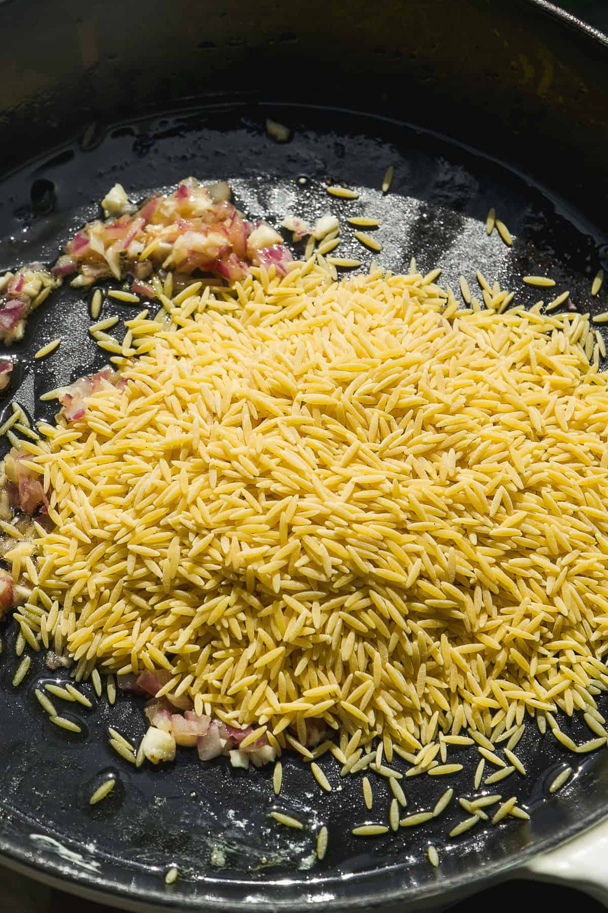 A close-up of corn orzo pasta and chopped onions in a black skillet, with the onions slightly sautéed and the orzo uncooked.