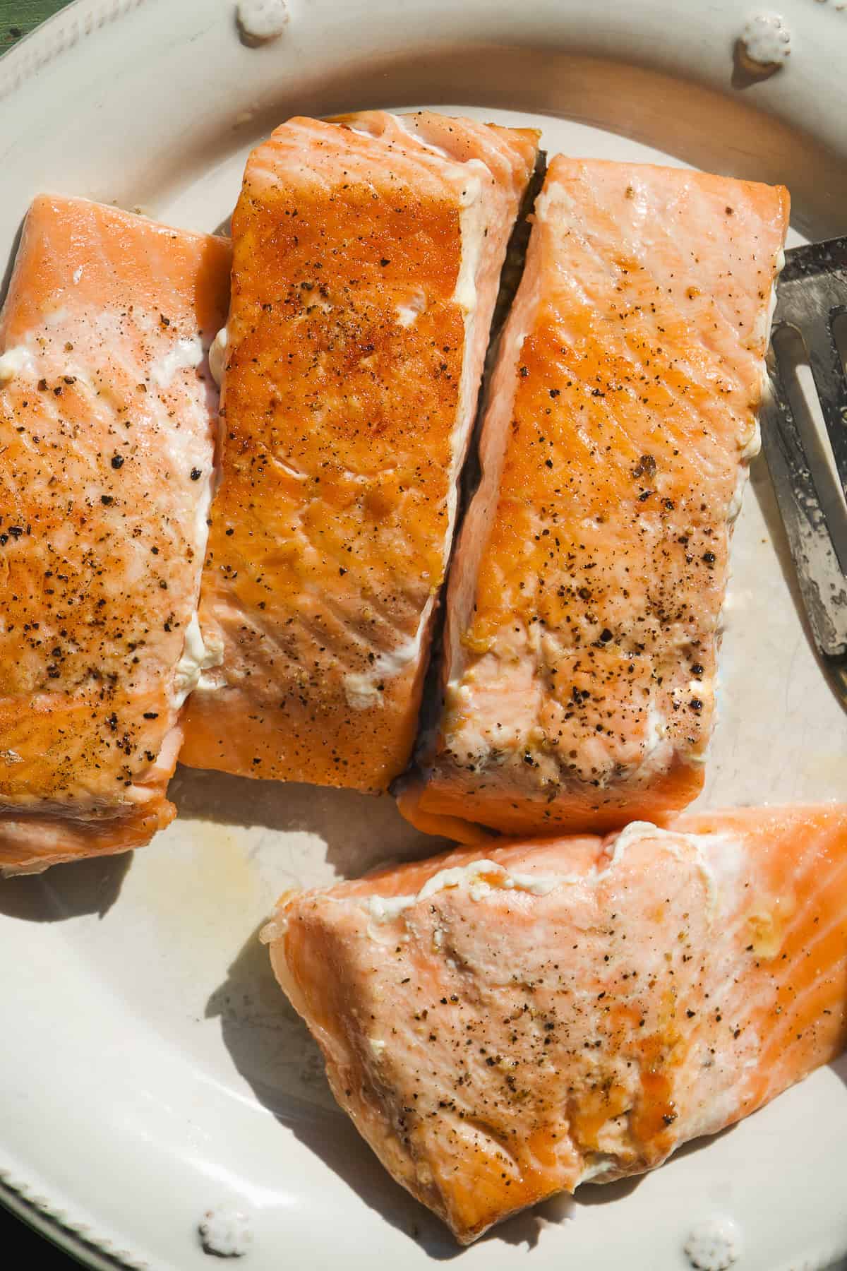Four cooked salmon fillets seasoned with pepper are arranged on a white plate alongside a flavorful corn orzo. A serving utensil is visible on the right side of the plate, and the salmon appears flaky and moist.