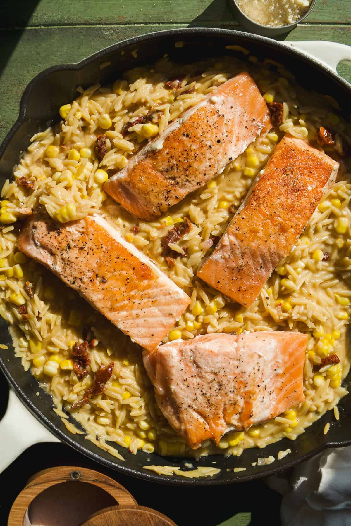 Four seared salmon fillets rest on a bed of creamy corn orzo pasta with sun-dried tomatoes in a skillet. The dish is garnished with pepper and displayed on a green wooden surface.