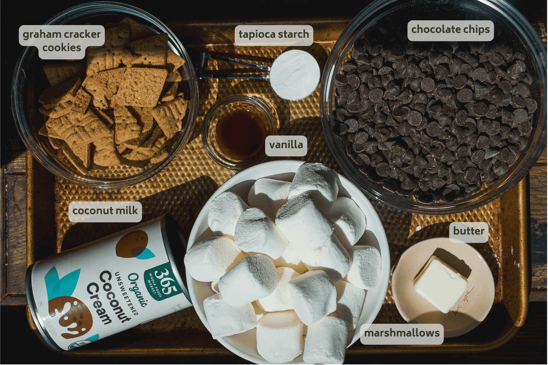A tray with labeled ingredients in bowls for smore pie: graham cracker cookies, chocolate chips, tapioca starch, vanilla, coconut milk, coconut cream, butter, and marshmallows.
