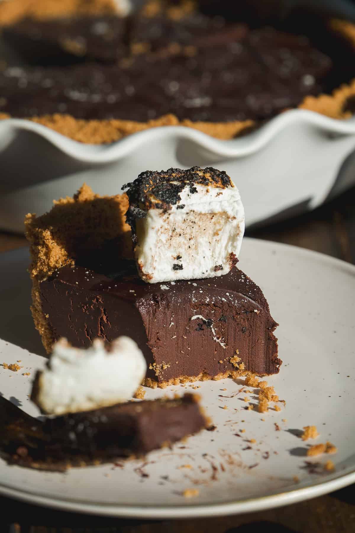 A slice of rich smore pie with a graham cracker crust sits on a plate, topped with a toasted marshmallow. More chocolate pie and crust are visible in a white pie dish in the background.