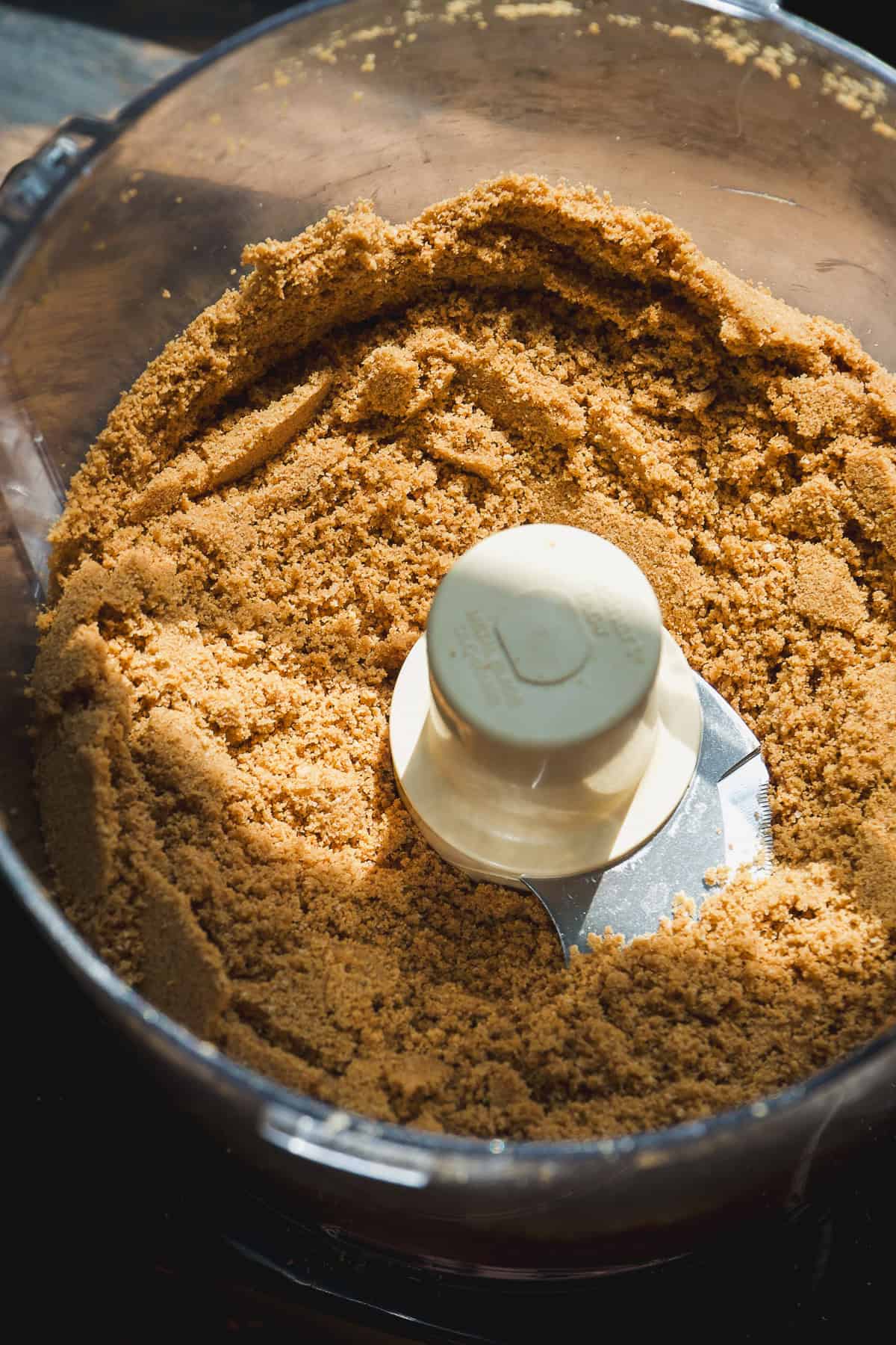 A close-up of a food processor bowl filled with finely ground brown crumbs, likely graham crackers or similar for a smore pie, with the metal blade in the center.
