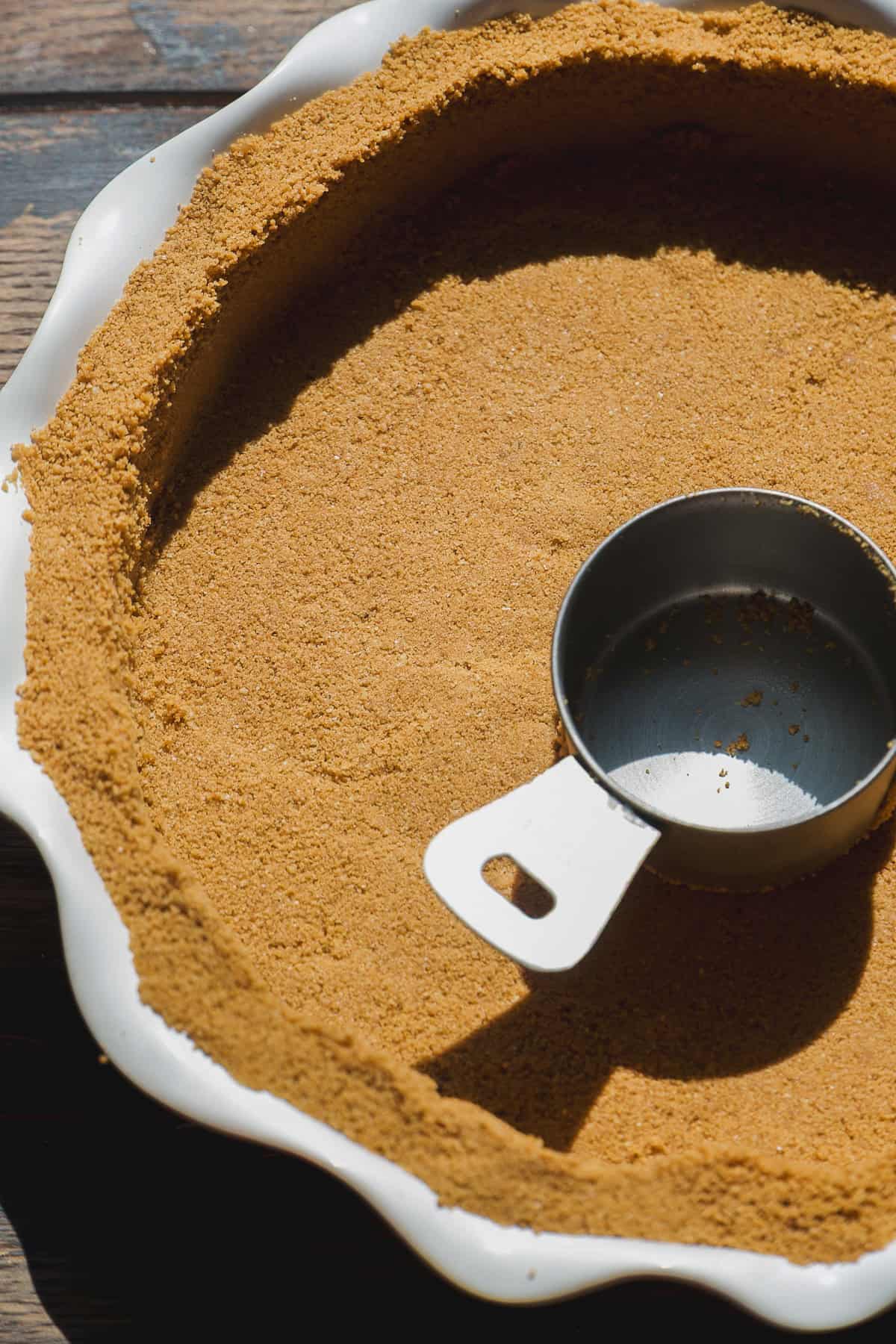 A metal measuring cup presses down a graham cracker crust in a white pie dish on a wooden surface, forming an even base for a delicious smore pie.
