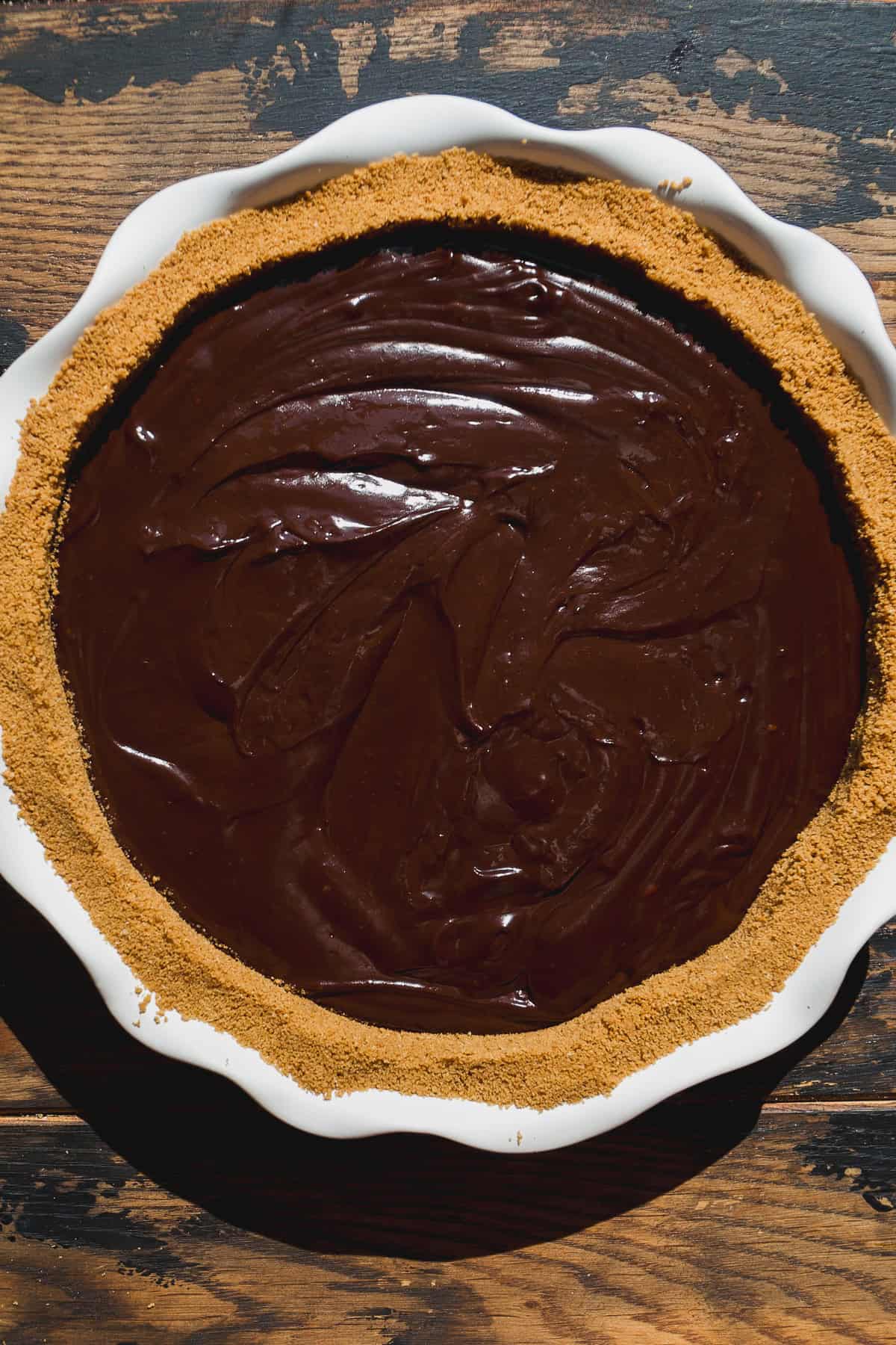 A pie dish filled with a smooth, glossy layer of chocolate filling, surrounded by a crumbly graham cracker crust, this smore pie sits on a rustic wooden surface.