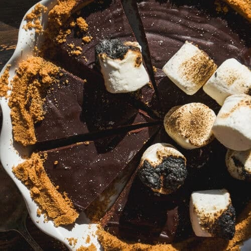 A smore pie with a crumb crust is sliced and topped with toasted and untoasted marshmallows in a white pie dish on a wooden surface. Some pie crumbs and a pie server are visible nearby.