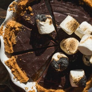 A smore pie with a crumbly crust in a white dish, topped with toasted and untoasted marshmallows. A few slices are missing, and crumbs are scattered around the pie on a wooden surface.