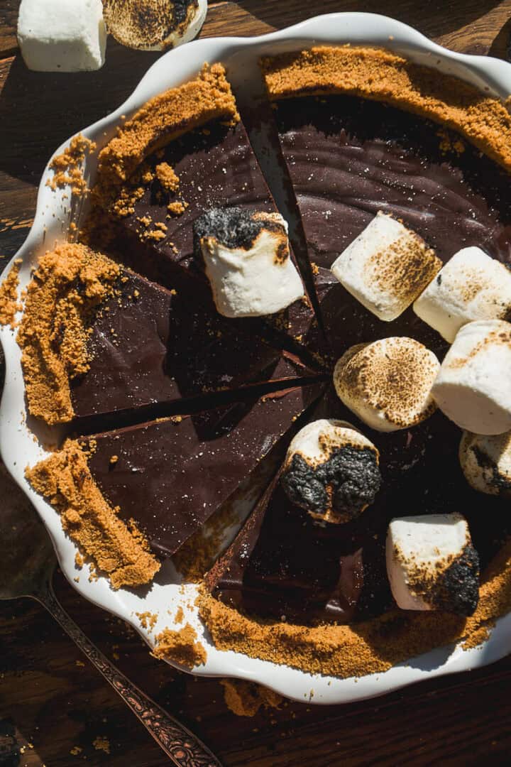 A smore pie with a crumb crust is sliced and topped with toasted and untoasted marshmallows in a white pie dish on a wooden surface. Some pie crumbs and a pie server are visible nearby.